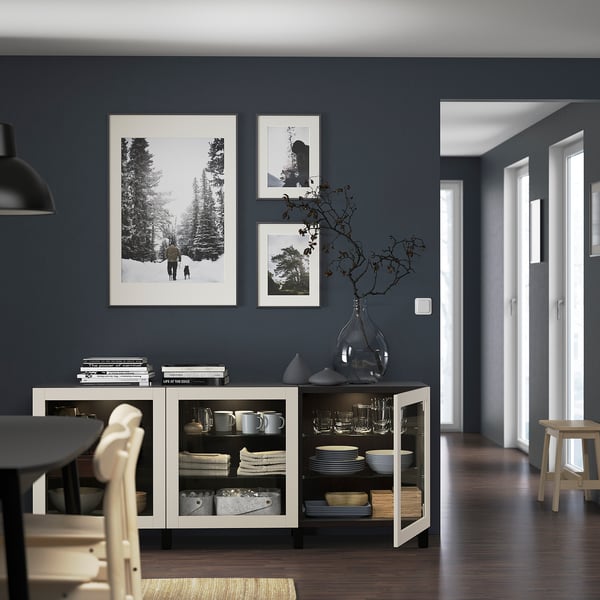 Sala de jantar moderna com aparador BESTÅ branco com pratos, canecas e artigos decorativos. Fotos em preto e branco adornam a parede cinza escuro.