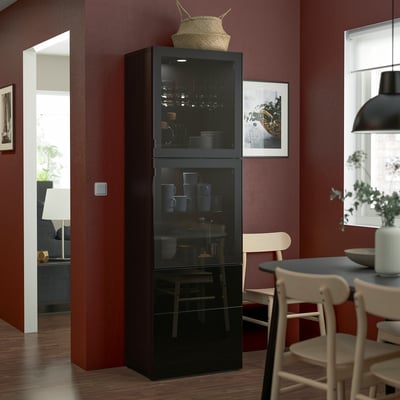 Armário preto BESTÅ com portas de vidro mostrando copos e pratos, fino entre cadeiras em uma sala vermelha, coberto com uma cesta e imagem.