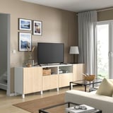 Sala de estar moderna com suporte de TV de madeira BESTÅ em cores claras, exibindo uma TV de tela plana, fotos emolduradas, luminária e vaso na parte superior.