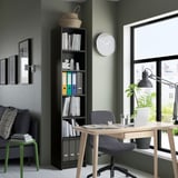 Estante preta de BILLY embalada com livros, ao lado da janela e da cadeira. Design alto e funcional com aglomerado.