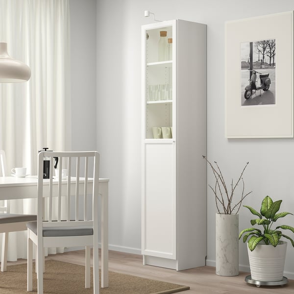Estante BILLY branca com porta de vidro e painel, design moderno, alta. Ao lado de uma cadeira branca, encaixada com mesa de jantar e planta grande.