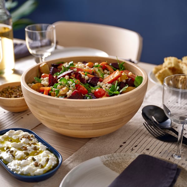 Tigela de madeira cheia de salada colorida fica na mesa perto de recipientes menores com laterais e copos.