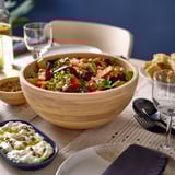 Tigela de madeira cheia de salada colorida fica na mesa perto de recipientes menores com laterais e copos.