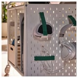 Unidade de armazenamento de jogos branca com pegboard, segurando fones de ouvido, alças e cabos. Rodas para mobilidade, projetadas para organizar equipamentos de jogos.
