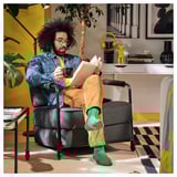 Homem de jaqueta jeans senta-se na versátil espreguiçadeira BRÄNNBOLL, segurando café e lendo, mostrando seu conforto e mobilidade.