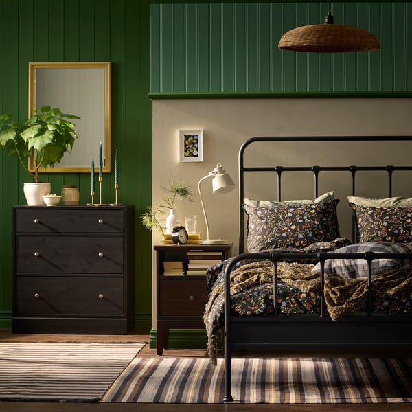 Quarto verde acolhedor com roupa de cama floral, cómoda decorada, candeeiro, plantas e tapete às riscas.