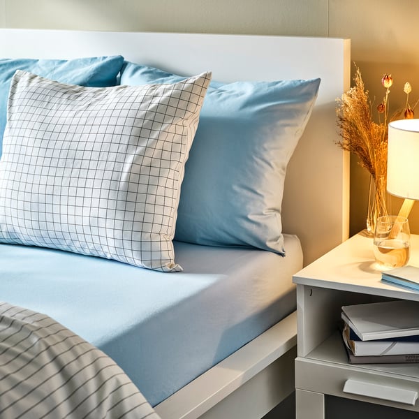 Lençol azul de encaixe na cama com almofada aos quadrados. 100% algodão, colchão grosso, livros de mesa de cabeceira e candeeiro. Ambiente acolhedor e convidativo para dormir.