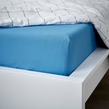 Uma cama bem arrumada com lençol azul e capa estampada branca, exibindo suas bordas elásticas.