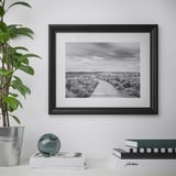 Uma foto costeira em preto e branco emoldurada está pendurada na parede, com um vaso de plantas e livros embaixo.