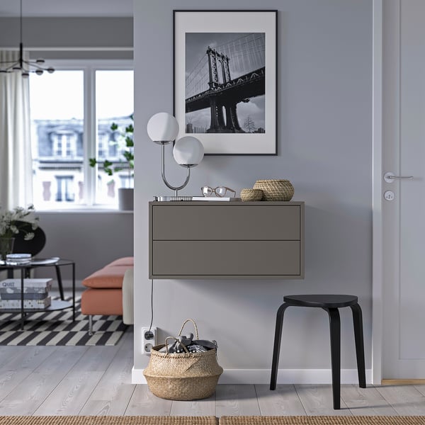 Quarto moderno: arte de ponte emoldurada em preto, luminária de parede, armário cinza com gavetas, banco laranja, banquinho preto, cesta grande com chaves.