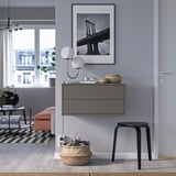 Quarto moderno: arte de ponte emoldurada em preto, luminária de parede, armário cinza com gavetas, banco laranja, banquinho preto, cesta grande com chaves.