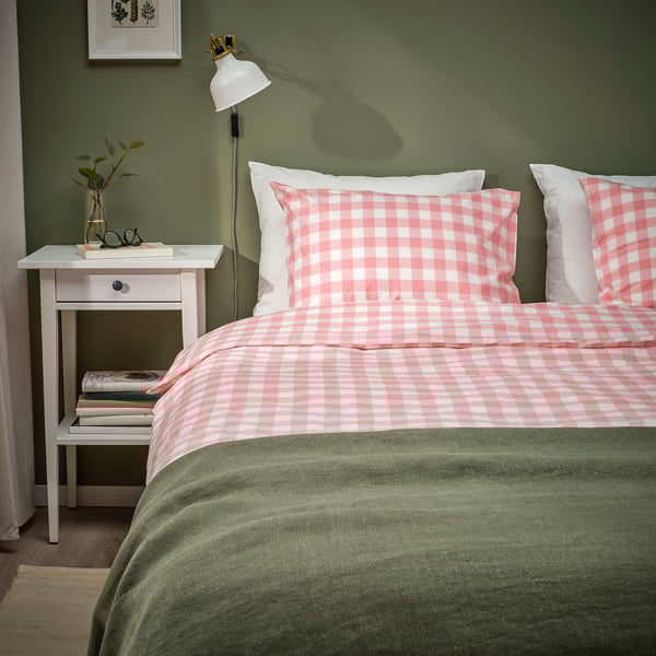 Quarto aconchegante com cama xadrez rosa, detalhes em verde e mesa de cabeceira com lâmpada e livros.