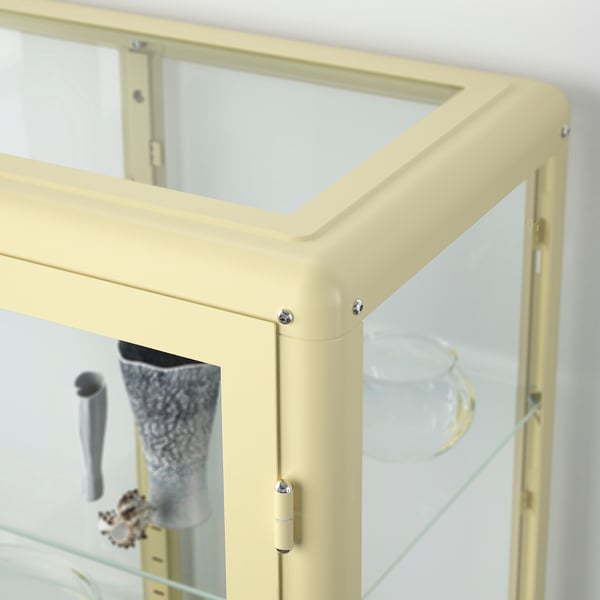 Um armário decorativo com porta de vidro de cor creme, oferecendo armazenamento versátil com prateleiras ajustáveis, garantindo estabilidade com pés ajustáveis.
