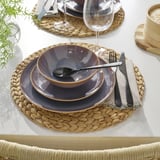 Mesa com jogo americano tecido, pratos rústicos, copos, talheres e guardanapo de pano.