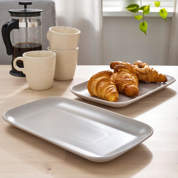 Mesa com três croissants em prato retangular, duas canecas empilhadas e uma prensa de café.