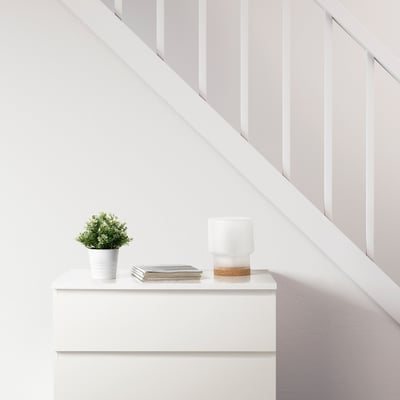 Sala minimalista com gaveta branca, planta FEJKA, revistas e candeeiro com base de cortiça.