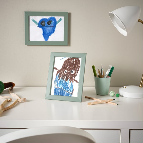 Dois desenhos infantis em molduras azuis claras: um yoda, uma pessoa. As molduras sentam-se na mesa branca com materiais de arte.
