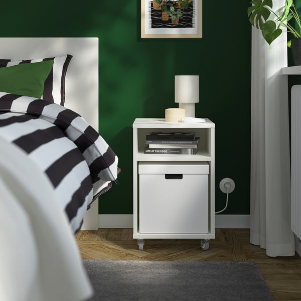 Quarto moderno com paredes verdes, mesa de cabeceira branca ao lado da cama, candeeiro, livros e plantas, com caixa FJÄDERHARV para arrumação.