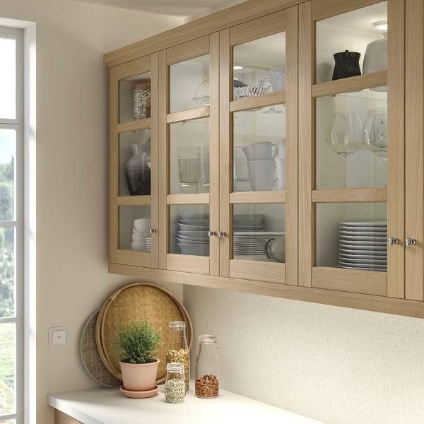 Armários de cozinha de madeira com portas de vidro, exibindo pratos, xícaras, taças de vinho. Abaixo, um balcão branco contém uma bandeja, planta, potes. Design clássico.