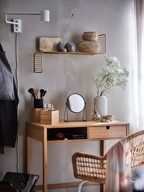 Vaidade minimalista: mesa de madeira, espelho, ferramentas de maquiagem, flores, cadeira de vime, prateleira de parede com cestos e decoração.