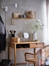 Vaidade minimalista: mesa de madeira, espelho, ferramentas de maquiagem, flores, cadeira de vime, prateleira de parede com cestos e decoração.