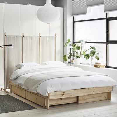 Quarto minimalista com uma cama extensível GLAMBERGET de madeira clara, lençóis brancos e uma grande janela com plantas.
