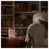Vela perfumada GLANSLIND acesa em vidro sobre mesa de madeira ao lado de poltrona, caneca, por estante.