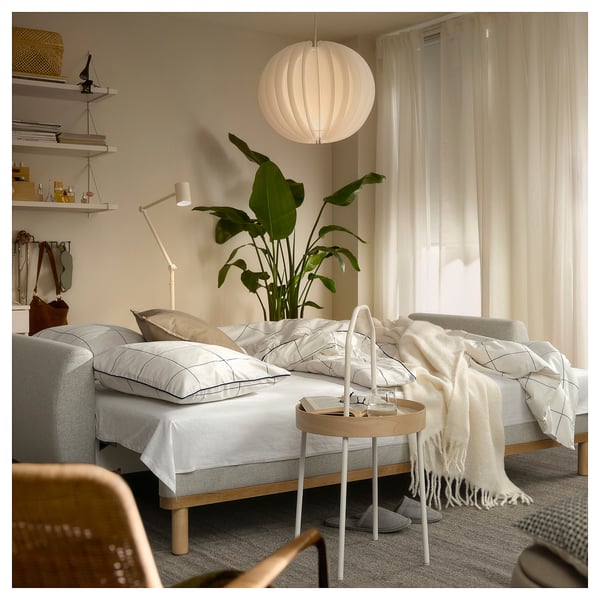 Sala de estar moderna com sofá-cama GRUNNARP, almofadas, cobertor, mesa de apoio, planta, candeeiro, estantes, cadeira, tapete e cortinas.