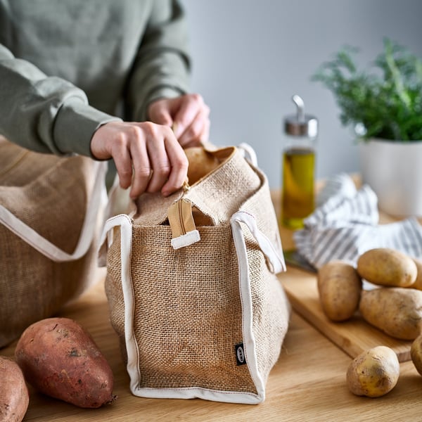 Pessoa segurando um saco de armazenamento GULLRISMOTT bege, cheio de batatas. Saco feito de juta natural, projetado para armazenar vegetais de raiz.