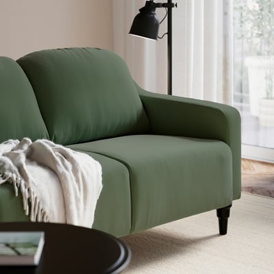 Sofá verde GULLVALLA com assento e encosto confortáveis, manta branca coberta, candeeiro preto ao lado, espaço de estar acolhedor.