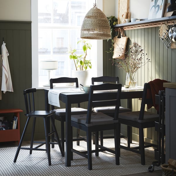 Conjunto de jantar aconchegante com mesa preta e cadeiras na janela. Conjunto de mesa com corredor, tigela e planta. Candeeiro suspenso no alto, prateleiras com decoração e planta.