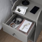 Mesa de cabeceira com gaveta aberta, mostrando fones de ouvido, um livro e um relógio. A prateleira superior contém um livro e um telefone. Cor cinza, design elegante.