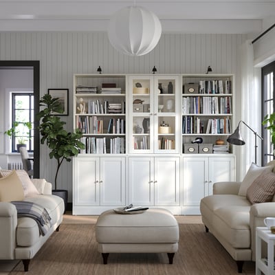 Elegante sala de estar com estantes de pinho branco HAVSTA, sofá, pufe, candeeiro, janela grande e plantas.