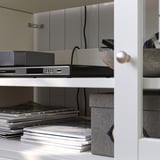 Prateleira de madeira branca com eletrônicos e revistas. Controle remoto e cabos pretos.