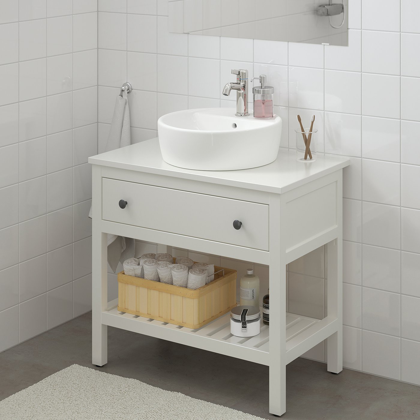 Pia HEMNES branca com pia redonda, gaveta e prateleira. A cesta contém papel higiênico e xampu, toalha cinza nas proximidades.
