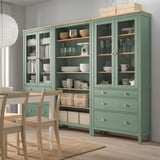 Módulo de arrumação verde HEMNES, portas de vidro, prateleiras de madeira, taças, louças, copos de vinho, livros, cadeiras.