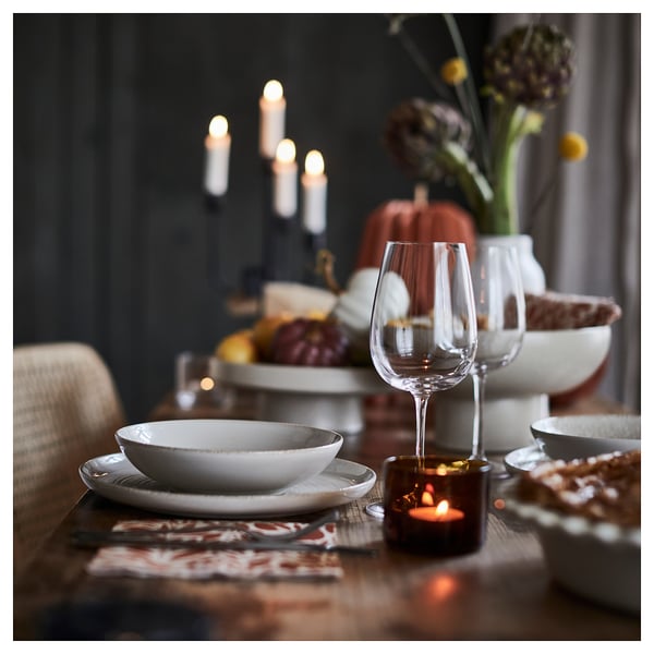 Elegante mesa de outono com pratos brancos, copo de vinho de vidro transparente e vela âmbar em suporte marrom. O pano de fundo escuro destaca decorações quentes.