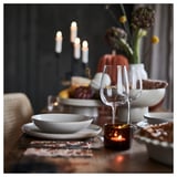 Elegante mesa de outono com pratos brancos, copo de vinho de vidro transparente e vela âmbar em suporte marrom. O pano de fundo escuro destaca decorações quentes.