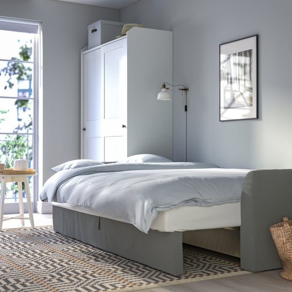 Quarto moderno com paredes azuis claras, uma cama branca com saia de cama cinza, mesa lateral de madeira, tapete estampado e janela com plantas.