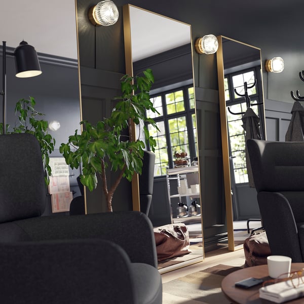 O grande espelho HOVET com moldura dourada domina a sala, refletindo plantas e móveis, adicionando profundidade.