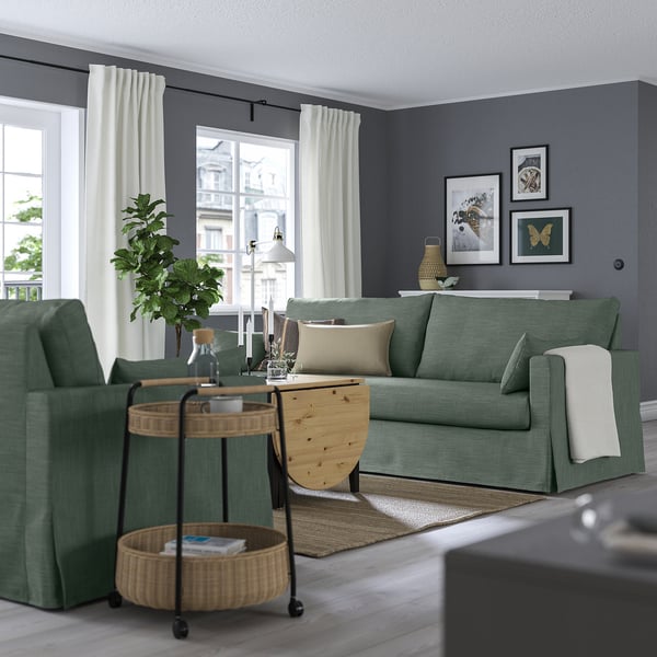 Sala de estar moderna com sofá verde sálvia HYLTARP, luz natural, planta e cobertor.