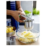 Pessoa usando prensa de batata de aço inoxidável para amassar batatas cozidas na tigela. Balcão de cozinha com limões, sal e ervas visíveis.