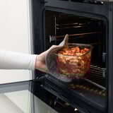 Pessoa colocando um recipiente de vidro com massa em um forno. O recipiente tem uma tampa de bambu, adequada para armazenamento de geladeira.