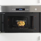 Micro-ondas embutido em aço inoxidável com recipiente de vidro no interior, exibindo sua função como aquecedor de alimentos.