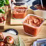 Recipientes cheios de molho de tomate, mostrando armazenamento e preservação. Tomates frescos e ervas nas proximidades.