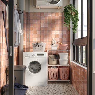 Lavanderia limpa com armazenamento JOSTEIN branco, lixeiras rosa, máquina de lavar, paredes de azulejos rosa e plantas penduradas perto da janela.