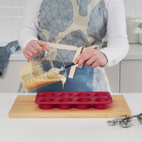 Uma pessoa despeja a massa em um molde de muffin de silicone vermelho colocado em uma tábua de madeira, demonstrando seu uso para assar muffins ou sobremesas.