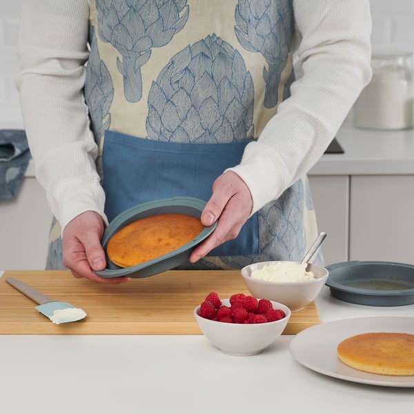 Pessoa segurando um molde redondo com um bolo assado dentro, ao lado de tigelas de creme e framboesas em um balcão de cozinha.