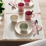 Mesa posta com canecas rosa, tigela verde, prato branco, colheres e guardanapos estampados em jogos americanos bege.