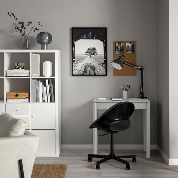Espaço de trabalho minimalista com secretária branca KJELLERIK, cadeira giratória preta, candeeiro e quadro de avisos entre a estante e o sofá.
